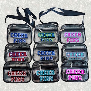 May include: Several clear, rectangular crossbody bags with black straps. Each bag displays the text "CHEER PINS" in different colours, such as blue, green, red, and pink. The bags are secured with a zip.