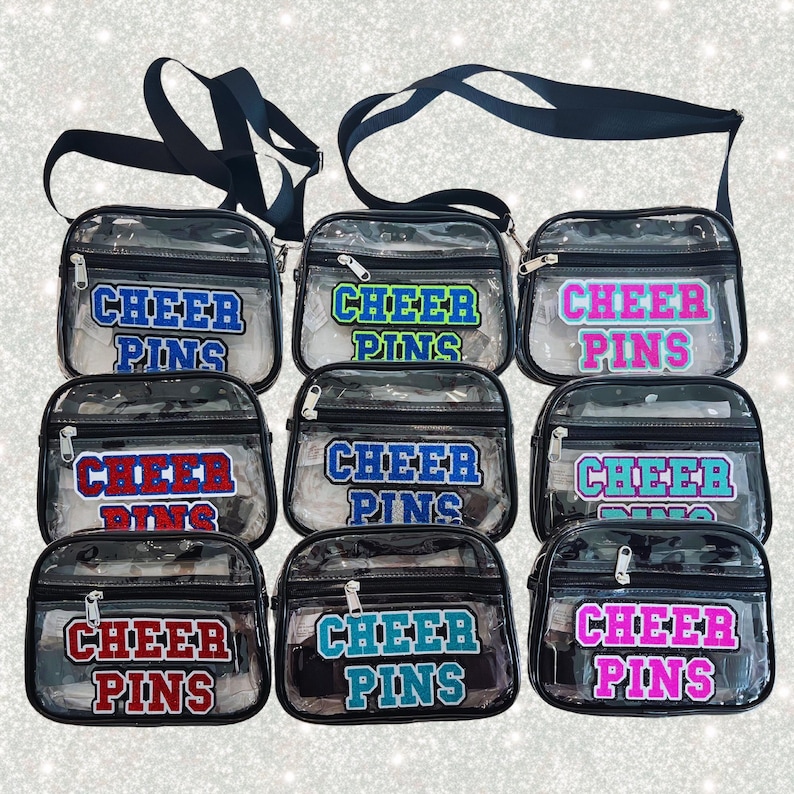 May include: Several clear, rectangular crossbody bags with black straps. Each bag displays the text "CHEER PINS" in different colours, such as blue, green, red, and pink. The bags are secured with a zip.