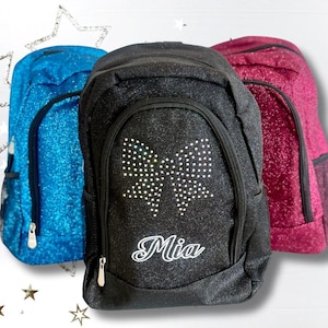 May include: Three glittery backpacks in blue, black, and magenta. The black backpack has a bow design made of small white dots and the name "Mia" in cursive. Each backpack has a zippered front pocket and side pocket.