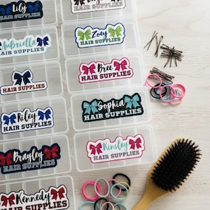 Könnte beinhalten: Eine Sammlung rechteckiger Aufbewahrungsboxen aus durchsichtigem Kunststoff mit Trennwänden, die jeweils mit einem personalisierten Etikett mit Namen und dem Text HAIR SUPPLIES versehen sind. Enthalten sind schwarze Haarnadeln, bunte Haargummis und eine Haarbürste.