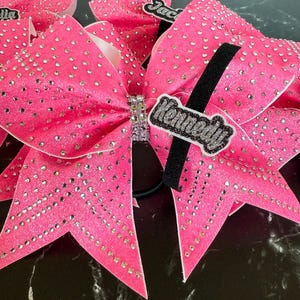 May include: A bright pink glittery hair bow adorned with numerous clear rhinestones. The center features a band of crystals, and a black fabric strip displays the name Kennedy in white glittery script lettering attached to the bow surface.