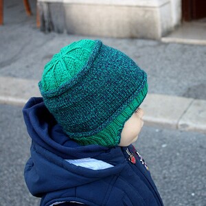 Puede incluir: Un niño con una chaqueta azul y un gorro de punto verde y azul con un borde doblado.