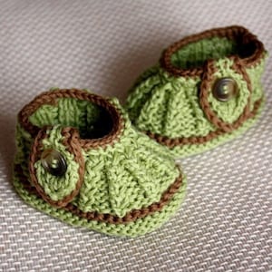 Puede incluir: Un par de botitas de bebé de punto verde con ribete marrón y un botón en cada botita.