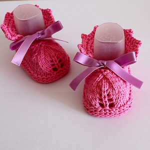 Puede incluir: Un par de botitas de bebé de punto rosa con lazos de satén morado. Las botitas tienen un delicado patrón de encaje y son perfectas para un recién nacido.