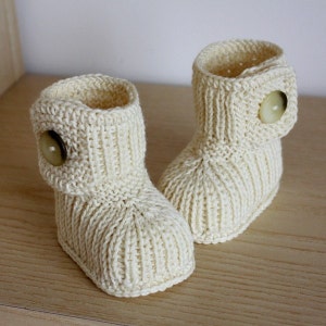 Könnte beinhalten: Ein Paar cremefarbene gestrickte Babyschuhe mit Knopfdetail am Bündchen.