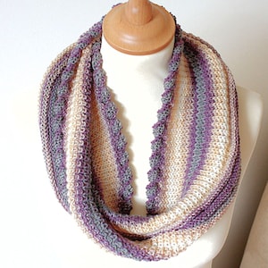 May include: A knitted infinity scarf featuring a striped pattern in shades of purple, grey, and cream. The scarf has a textured, ribbed knit construction with a decorative, scalloped edge detail running along the inner length of the loop.