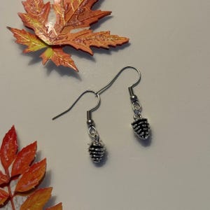 May include: Silver-coloured drop earrings featuring miniature pinecone charms. The earrings are set against a white backdrop, with decorative autumn leaves. These earrings are a nature-themed accessory.