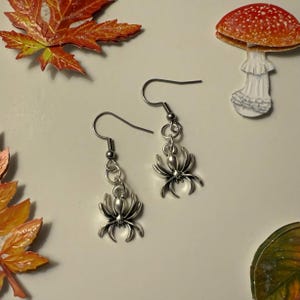 May include: A pair of silver spider earrings. Each earring has a spider charm hanging from a hook. The earrings are shown with autumn decorations, including fall leaves and a mushroom.