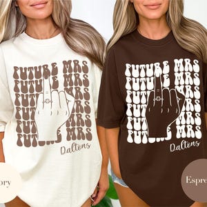 Puede incluir: Dos camisetas de gran tamaño con un diseño marrón y blanco. El diseño presenta el texto "FUTURE MRS" repetido varias veces con un dedo medio levantado y un anillo en el dedo. El texto "Daltons" está impreso debajo del diseño. Las camisetas se muestran en dos colores: marfil y espresso.