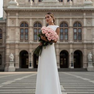 Könnte beinhalten: Eine Frau in einem weißen, bodenlangen Kleid hält einen Strauß rosa Rosen. Das Kleid hat ein schlichtes Design mit hohem Ausschnitt und kurzen Ärmeln. Der Hintergrund zeigt ein großes, verziertes Gebäude mit Bogenfenstern und Statuen.