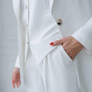 May include: A crisp, white suit featuring a blazer, vest, and trousers. The blazer has a single button closure and a notched lapel. The vest has a button-down front. The trousers have a pleated front and pockets. The suit is made of a smooth, wrinkle-resistant fabric.
