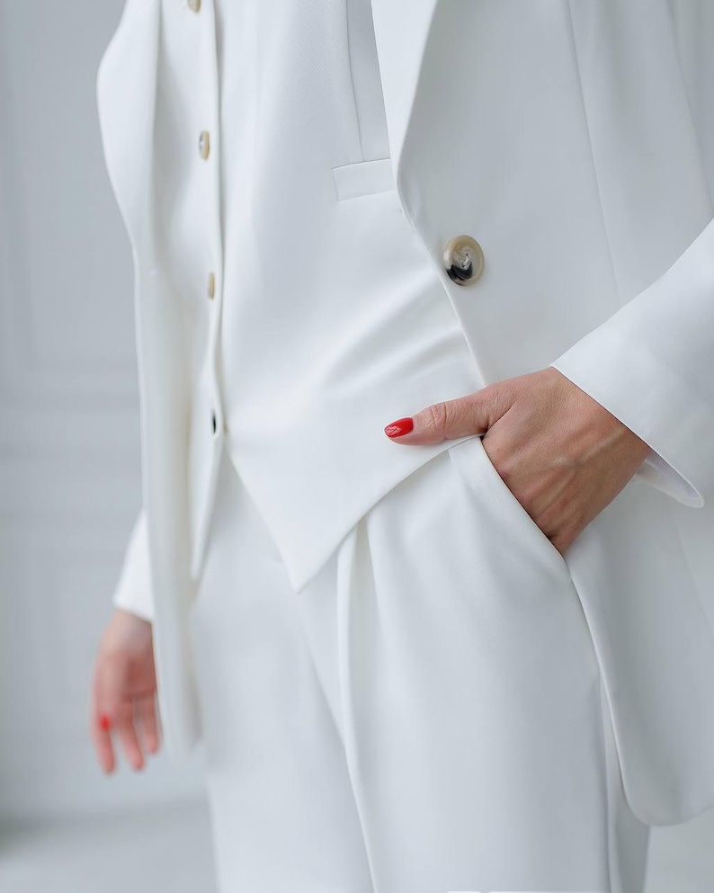 May include: A crisp, white suit featuring a blazer, vest, and trousers. The blazer has a single button closure and a notched lapel. The vest has a button-down front. The trousers have a pleated front and pockets. The suit is made of a smooth, wrinkle-resistant fabric.