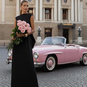 Könnte beinhalten: Eine Frau in einem schwarzen Kleid hält einen Strauß rosa Rosen und steht neben einem Oldtimer-Cabriolet in Rosa. Das Auto hat Weißwandreifen und einen cremefarbenen Innenraum. Im Hintergrund ein großes Gebäude mit Säulen.