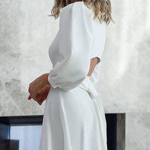 Pu&ograve; includere: Abito bianco con maniche lunghe a sbuffo e gonna fluida. L'abito ha la schiena scoperta e un fiocco in vita. Il tessuto sembra essere un materiale liscio e leggero. Adatto per occasioni formali.
