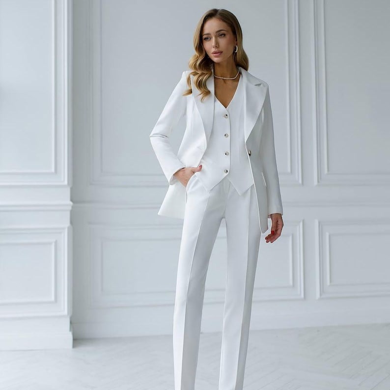 May include: A three-piece white suit consisting of a blazer, vest, and trousers. The blazer has a notched lapel and the vest has buttons. The trousers are straight-legged. The suit is made of a smooth, solid white fabric.