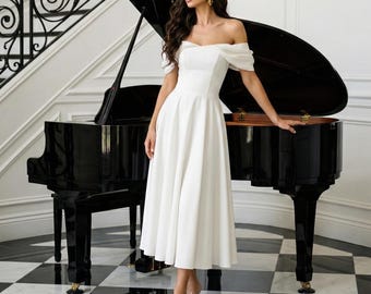 Può includere: Abito midi bianco con spalle scoperte, corpetto aderente e gonna svasata. L'abito è abbinato a tacchi nude. Un pianoforte a coda nero è sullo sfondo, su un pavimento a scacchi bianco e nero.