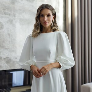 Pu&ograve; includere: Un abito bianco con scollatura a barchetta e maniche lunghe a sbuffo. L'abito ha una vita aderente e una gonna svasata. La donna indossa orecchini pendenti lunghi. Lo sfondo comprende un camino e tende.