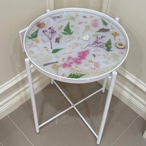 May include: White side table with a round, white top featuring pressed flowers and gold leaf accents. The table has a white metal frame with a cross-shaped base.