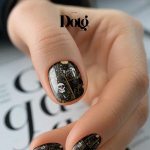 May include: A fingernail featuring a black and gold nail wrap design with a white skull graphic and celestial motifs. The nail art displays a detailed pattern of tarot-style cards with intricate gold linework and small white stars on a dark background.