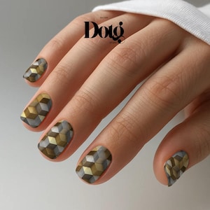 May include: Close-up of a hand with geometric nail art. The nails feature a 3D cube design in shades of gold, gray, and silver. The image includes the text "Dotg Studio" in a black, elegant font.