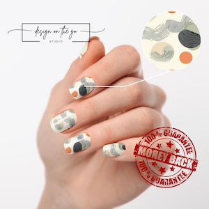May include: Nail wraps with an abstract design featuring gray, black, and orange shapes on a cream background. The nails are shown on a hand, with a close-up of the design in a circle. The text "design on the go studio" is in the upper left corner.