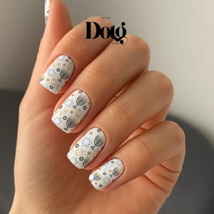 May include: White nail art featuring a repeating pattern of menorahs, Stars of David, and other Jewish symbols in gold, black, and light blue. The nails are on a hand with a neutral skin tone. The brand name "Dotg Studio" is visible in the background.