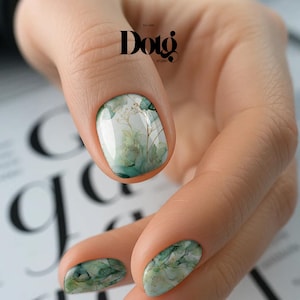 May include: Close-up of manicured nails with a watercolor design. The nails feature a base of white and green hues with gold accents, creating a botanical theme. The image includes the text "Doig Studio".