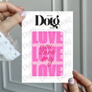 May include: A pink and white card with the text "LOVE YOU ARE MY LOVE" in a bold, blocky font. The card is held by a person with manicured nails. The top of the card reads "Doig Studio" with the text "SINCE 2025".