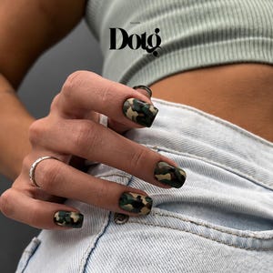 May include: Square-shaped fingernails featuring a matte camouflage pattern in shades of dark green, brown, black, and tan. The hand is positioned against light blue denim fabric, with a thin silver ring worn on the ring finger and a logo reading Dotg.