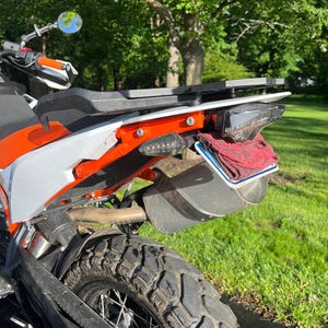 May include: Rear view of an orange and white motorcycle with a black tyre. The motorcycle has a black seat, a number plate, and a red cloth. The background is a green lawn and trees.