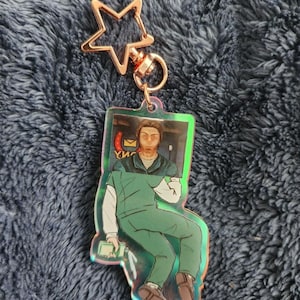 May include: A holographic acrylic keychain featuring a seated figure in green scrubs. The keychain has a star-shaped clasp in rose gold. The figure is holding a small device and a piece of paper. The background is a blurry image of a room.