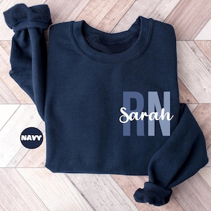 May include: Navy blue crewneck sweatshirt with the word "Sarah" in white script and the letters "RN" in blue. A navy blue circle with the word "NAVY" in white is in the lower left corner.