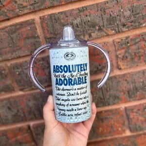 May include: White and blue insulated tumbler with two handles and a clear lid. The tumbler has a quote that reads "Absolutely Until the Next Feeding Adorable. This charmer's a master of cuteness. Don't be fooled! That angelic coo turns into a symphony of screams when the tummy needs a tune-up. Cuddle now, refuel later."