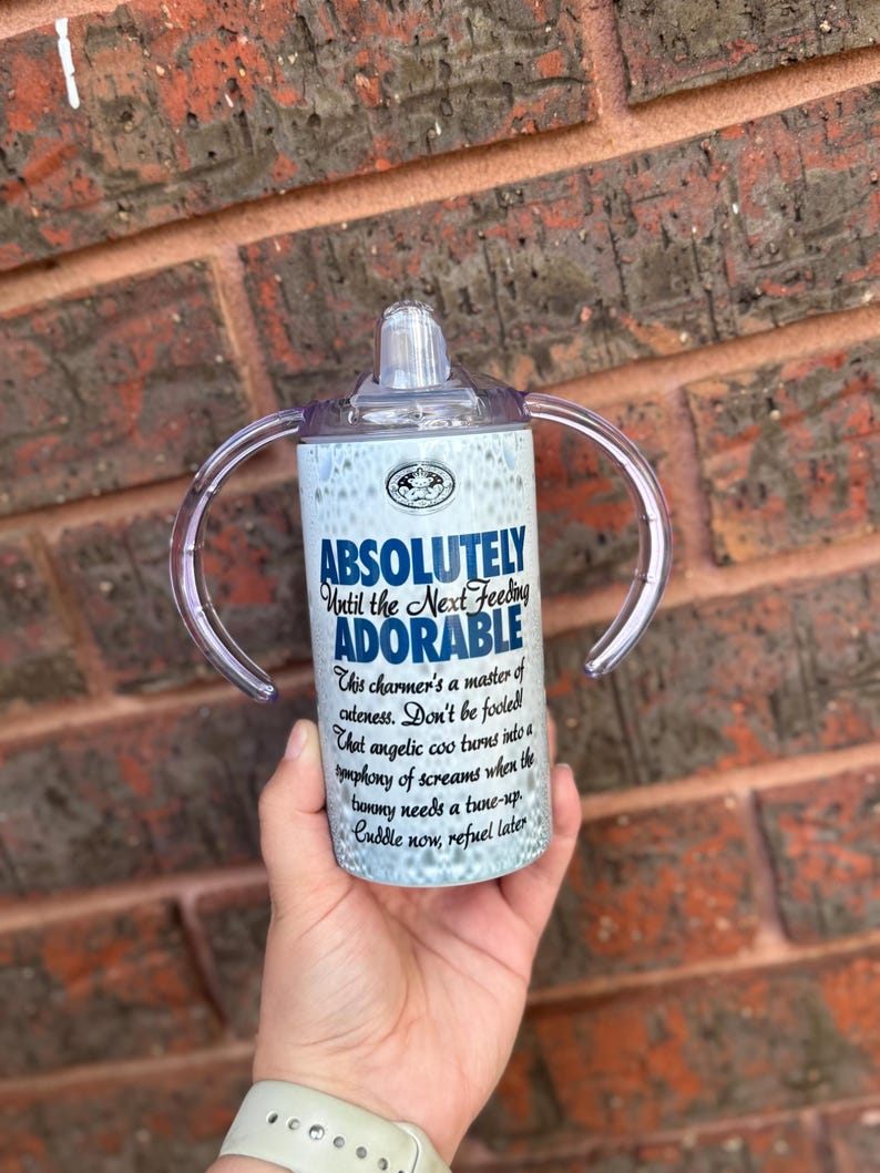 May include: White and blue insulated tumbler with two handles and a clear lid. The tumbler has a quote that reads "Absolutely Until the Next Feeding Adorable. This charmer's a master of cuteness. Don't be fooled! That angelic coo turns into a symphony of screams when the tummy needs a tune-up. Cuddle now, refuel later."