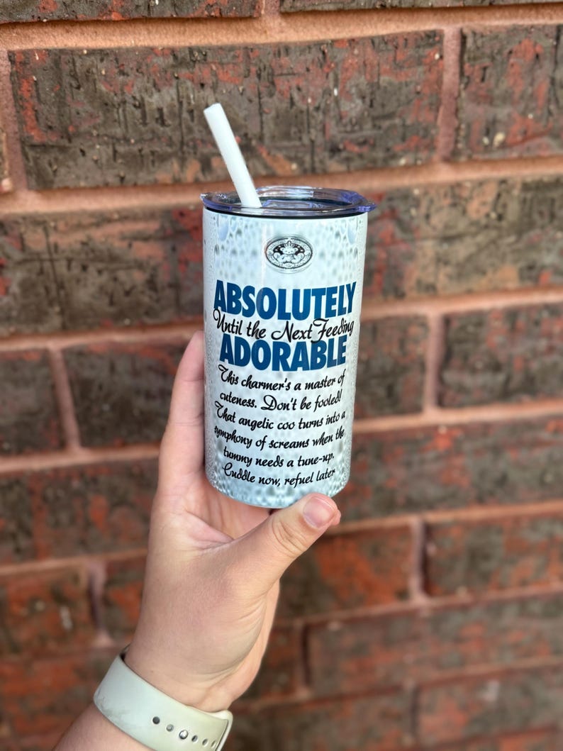 May include: White tumbler with a blue and black design that says "Absolutely Until the Next Feeding Adorable". The tumbler has a white straw and is being held by a hand.