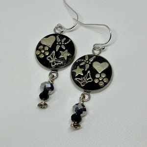 Puede incluir: Un par de pendientes colgantes de plata con dijes redondos en blanco y negro que representan una libélula, una mariposa, un corazón y una estrella. Cada dije está unido a una cuenta de cristal negro.