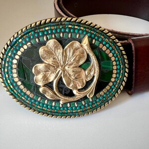 Puede incluir: Hebilla de cinturón ovalada con un diseño de trébol dorado. La hebilla tiene un borde de mosaico de cuentas turquesas y doradas, sobre un fondo verde oscuro. Se adjunta un cinturón de cuero marrón.