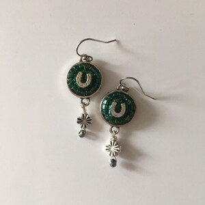 May include: A pair of dangle earrings featuring a circular design with a green mosaic background and a silver horseshoe. Each earring has a silver charm with a cross design and a small gray bead. The earrings have silver hooks.