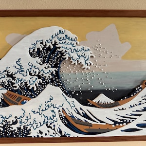 May include: A framed 3D artwork depicting a large wave crashing with a mountain in the background. The wave is painted in shades of blue, white, and black, with a traditional Japanese design. The mountain is painted in shades of gray and white. The artwork is framed in a brown wooden frame.