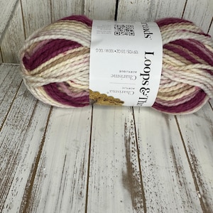 May include: A ball of Loops & Threads Charisma acrylic yarn in a mix of cream, beige, and burgundy shades. The label indicates 100 meters and 100 grams. The yarn is displayed on a white wooden surface.