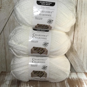 Puede incluir: Tres ovillos de lana blanca Charisma apilados sobre una superficie de madera. Cada ovillo tiene una etiqueta que dice "Charisma" y "Loops & Threads". Las etiquetas también indican "Patrón incluido" y "100 m / 100 g."