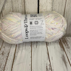 Puede incluir: Una madeja de hilo Loops & Threads Soft Rainbow en un esquema de color blanco y pastel. El hilo es 79% acrílico y 21% poliéster. La etiqueta indica una aguja de tamaño 7 (5 mm) y un calibre de 20 puntos. El hilo se lava a mano con agua fría, no se blanquea, se seca en plano y no se plancha.
