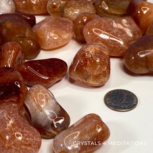 May include: A collection of polished carnelian stones in shades of orange and brown. The stones vary in size and shape, with a smooth, glossy finish. A US quarter coin is included for size reference. The text "CRYSTALS 4 MEDITATIONS" is visible.