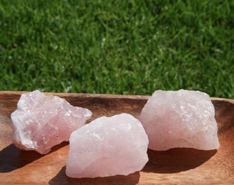 May include: Three pink rose quartz crystals on a wooden surface. The crystals are rough and have a natural, unpolished look.