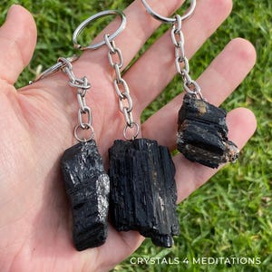 May include: Three black tourmaline crystal keychains with silver-colored chains and key rings. The raw, natural crystals vary in shape and size, held against a background of green grass. The text "CRYSTALS 4 MEDITATIONS" is visible at the bottom.