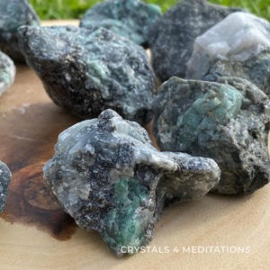 May include: A collection of rough emerald crystals, showcasing a mix of dark green and gray hues. The crystals are displayed on a wooden surface, with the text "CRYSTALS 4 MEDITATIONS" visible at the bottom. The natural stones vary in size and shape.