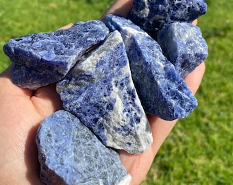 May include: A handful of rough-cut sodalite stones in varying sizes and shapes. The stones are a deep blue color with white and gray marbling. The image is taken outdoors with green grass in the background. The text "CRYSTALS 4 MEDITATIONS" is visible.