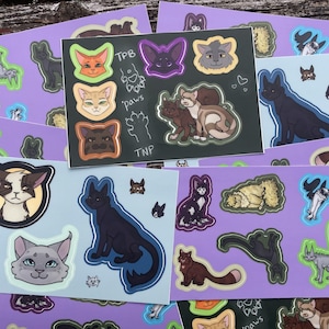 May include: A selection of cat stickers on purple and light blue cards. The stickers depict various cat designs in different colours, including black, brown, orange, and grey. Some stickers have text, including "TPB", "paws", and "TNP".