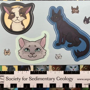May include: A sheet of cat-themed stickers featuring various cat illustrations. The stickers include a brown and white cat in a circular frame, a black cat, and several smaller cat faces. The stickers are on a light blue background.