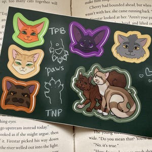 May include: A dark green sticker sheet with cat illustrations in orange, brown, grey, and purple. The sheet includes paw prints and the text "TPB", "paws", and "TNP".